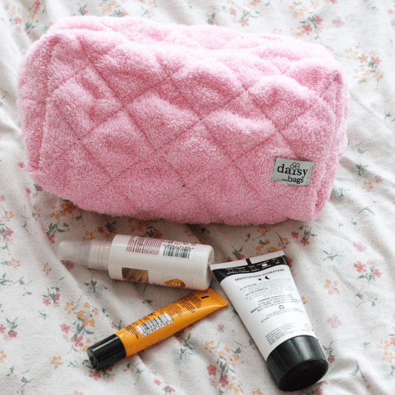 The Leela pink makeup bag on a floral bed sheet with 3 skincare products