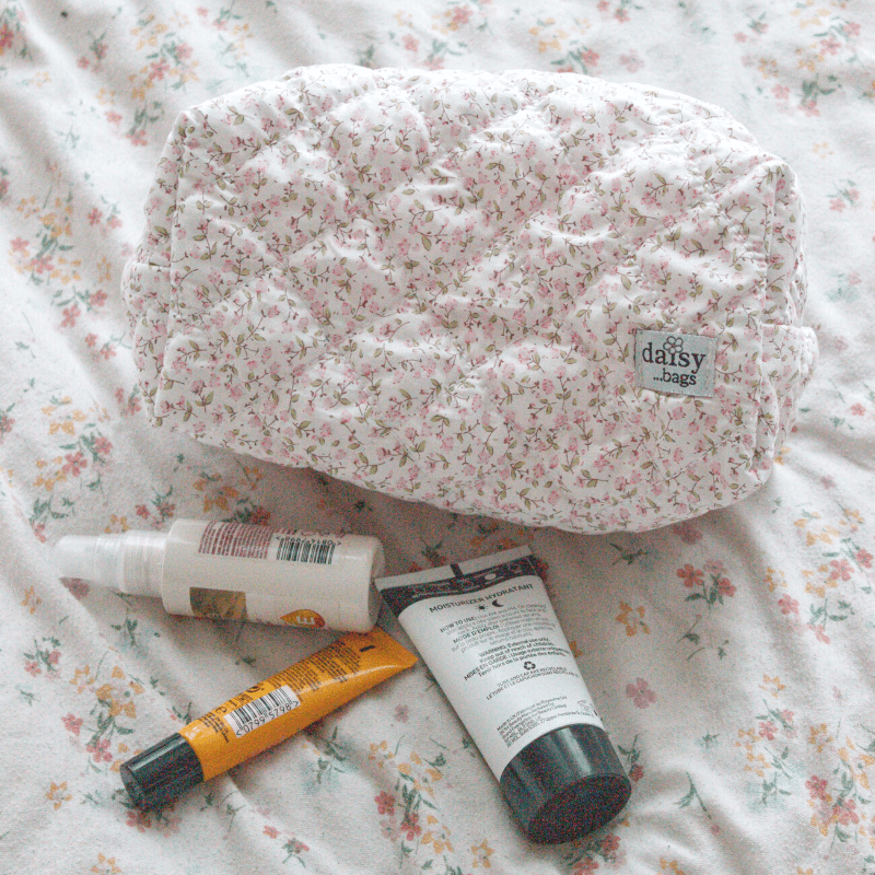 The Daisy floral makeup bag on a floral bed sheet with 3 skincare products